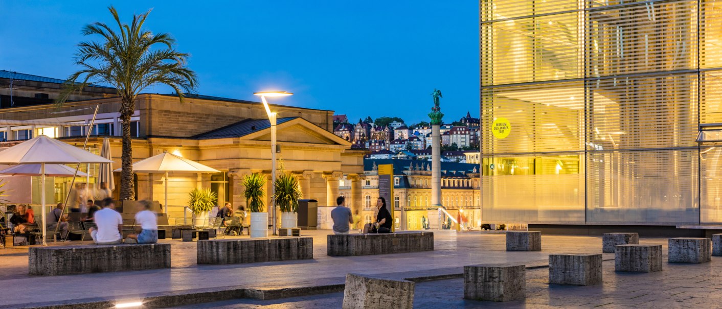 Evening atmosphere at Kleiner Schlossplatz with palm trees, people and modern architecture. Illuminated buildings and a statue can be seen in the background., © SMG, Werner Dieterich Evening atmosphere at Kleiner Schlossplatz with palm trees, people and modern architecture. Illuminated buildings and a statue can be seen in the background., © SMG, Werner Dieterich