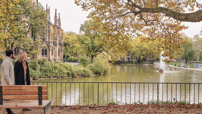 Ein Paar spaziert am Feuersee entlang, im Hintergrund die Johanneskirche und herbstliche Bäume. Ein Brunnen sprudelt im See., © SMG Christoph Düpper