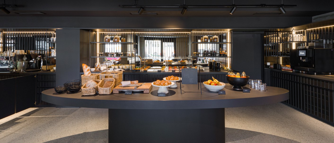Ein stilvolles Frühstücksbuffet mit Brotkörben, Obstschalen und Kaffeemaschine in einem modernen Raum., © EmiLu Hotel GmbH Ein stilvolles Frühstücksbuffet mit Brotkörben, Obstschalen und Kaffeemaschine in einem modernen Raum., © EmiLu Hotel GmbH
