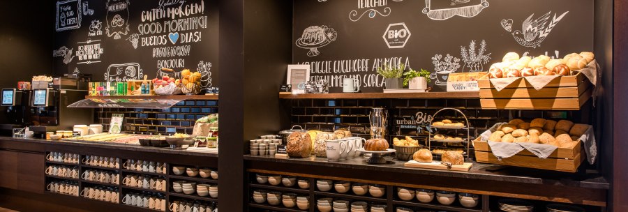 Ein Frühstücksbuffet mit Brotkörben, Tassen und Schüsseln vor einer dekorativen Kreidetafelwand., © TOMAS Ein Frühstücksbuffet mit Brotkörben, Tassen und Schüsseln vor einer dekorativen Kreidetafelwand., © TOMAS