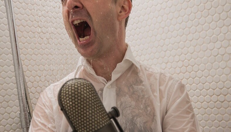 Ein Mann in einem wei&szlig;en Hemd singt leidenschaftlich in einer Dusche, w&auml;hrend er ein Mikrofon h&auml;lt. Wasser str&ouml;mt &uuml;ber ihn., &copy; Rosenau Kultur e.V.
