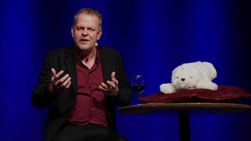 Ein Mann spricht auf einer B&uuml;hne, neben ihm ein Pl&uuml;schtier auf einem Tisch mit einem Glas Wein. Der Hintergrund ist blau beleuchtet., &copy; Theaterhaus Stuttgart e.V.