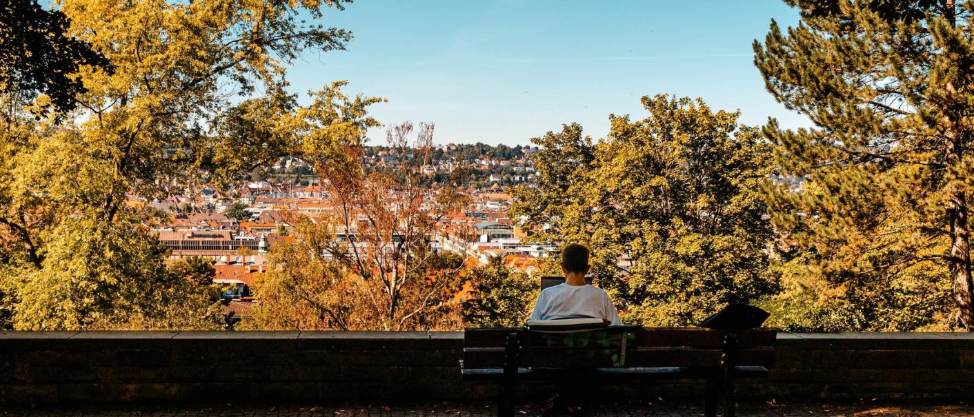 Eine Person sitzt auf einer Bank und schaut auf eine Stadt, umgeben von herbstlichen Bäumen. Der Himmel ist klar und blau., © Stuttgart Marketing GmbH, Sarah Schmid
