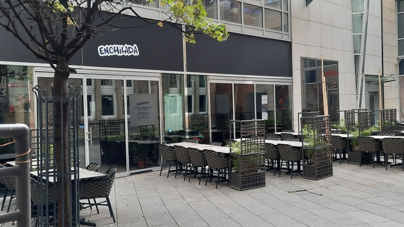 Outdoor area of a restaurant called Enchilada with empty tables and chairs. A tree and plants separate the seating areas., © SMG Outdoor area of a restaurant called Enchilada with empty tables and chairs. A tree and plants separate the seating areas., © SMG