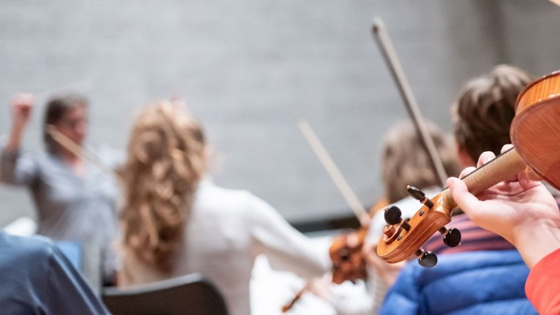 Ein Streichorchester probt, im Vordergrund ist eine Geige zu sehen. Der Dirigent hebt die Arme, Musiker sind unscharf im Hintergrund., © Jens Volle Ein Streichorchester probt, im Vordergrund ist eine Geige zu sehen. Der Dirigent hebt die Arme, Musiker sind unscharf im Hintergrund., © Jens Volle
