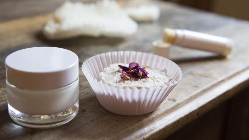 Ein Glas mit Creme, ein festes Kosmetikprodukt in einem Papierförmchen und ein Lippenpflegestift auf einem Holztisch., © hoponopo GmbH Ein Glas mit Creme, ein festes Kosmetikprodukt in einem Papierförmchen und ein Lippenpflegestift auf einem Holztisch., © hoponopo GmbH
