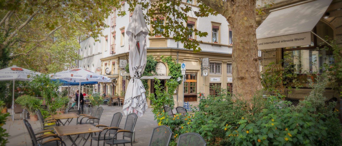 Ein Bistro am Wilhelmsplatz mit leeren Tischen und St&uuml;hlen im Freien, umgeben von gr&uuml;nen Pflanzen und B&auml;umen., &copy; Martina Denker