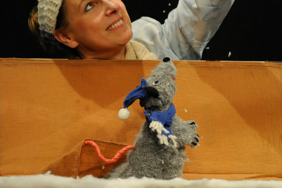 Eine Frau lächelt auf eine graue Mausfigur mit blauer Mütze und Schal, die aus einer Kiste schaut. Die Szene wirkt winterlich., © TheaterFusion