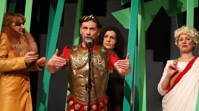 Vier Schauspieler in historischen Kostümen auf einer Bühne. Einer trägt eine römische Rüstung, die anderen sind in antiker und moderner Kleidung gekleidet., © Sabine Haymann