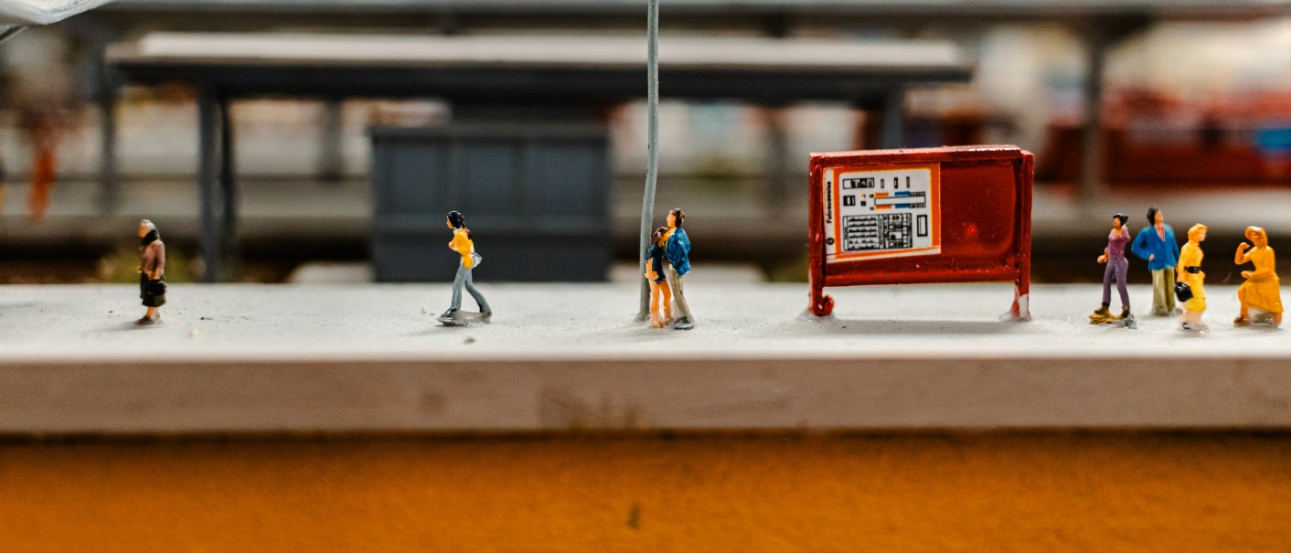 Miniaturfiguren an einem Modellbahnhof, neben einer Laterne und einem Fahrplan. Menschen in verschiedenen Posen, Hintergrund unscharf., © SMG_Sarah Schmid