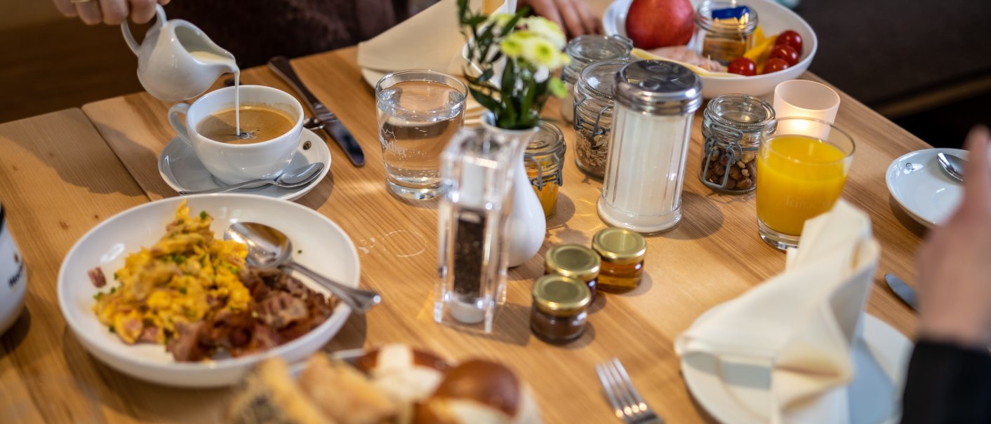 A laid breakfast table with coffee, scrambled eggs, bread rolls, orange juice and jam. One person pours milk into the coffee., &copy; TOMAS