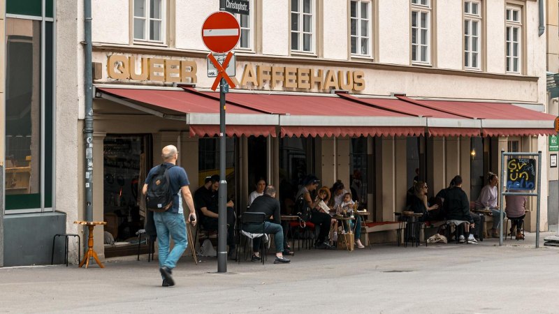 Ein Café mit rotem Vordach und dem Schriftzug 'Queer Kaffeehaus'. Menschen sitzen draußen an Tischen. Ein Mann geht vorbei., © SMG, Sarah Schmid Ein Café mit rotem Vordach und dem Schriftzug 'Queer Kaffeehaus'. Menschen sitzen draußen an Tischen. Ein Mann geht vorbei., © SMG, Sarah Schmid