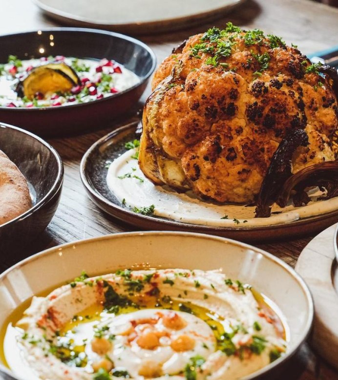 A table set with various Middle Eastern dishes, including roasted cauliflower, hummus with chickpeas, pita bread, and shakshuka with poached eggs in tomato sauce., © yafa, Stuttgart