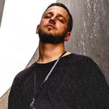 Ein Mann mit Bart und schwarzem Oberteil steht vor einer strukturierten Wand und blickt selbstbewusst in die Kamera., &copy; links im Bild