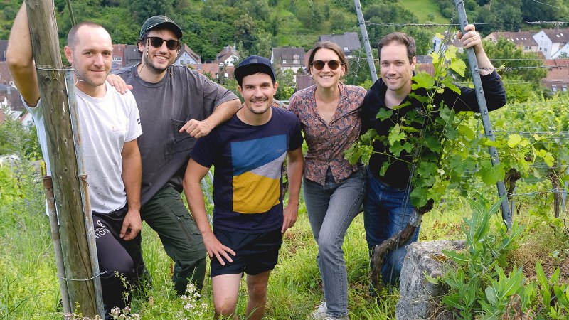 F&uuml;nf Personen posieren l&auml;chelnd in einem Weinberg. Im Hintergrund ist ein Dorf zu sehen. Die Gruppe wirkt entspannt und naturverbunden., &copy; Weinkollektiv Edenberg & Tal