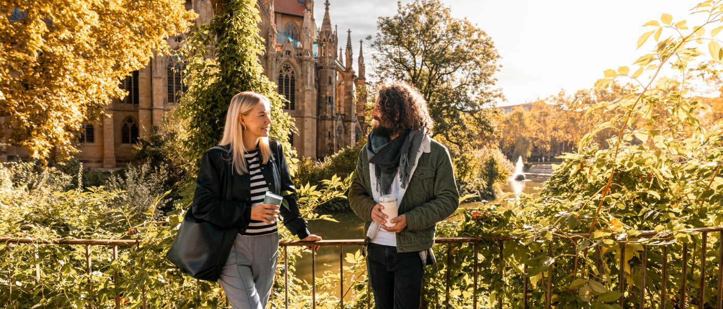 Zwei Personen unterhalten sich vor der Johanneskirche am Feuersee, umgeben von herbstlichem Laub und Sonnenschein., © SMG, Sarah Schmid