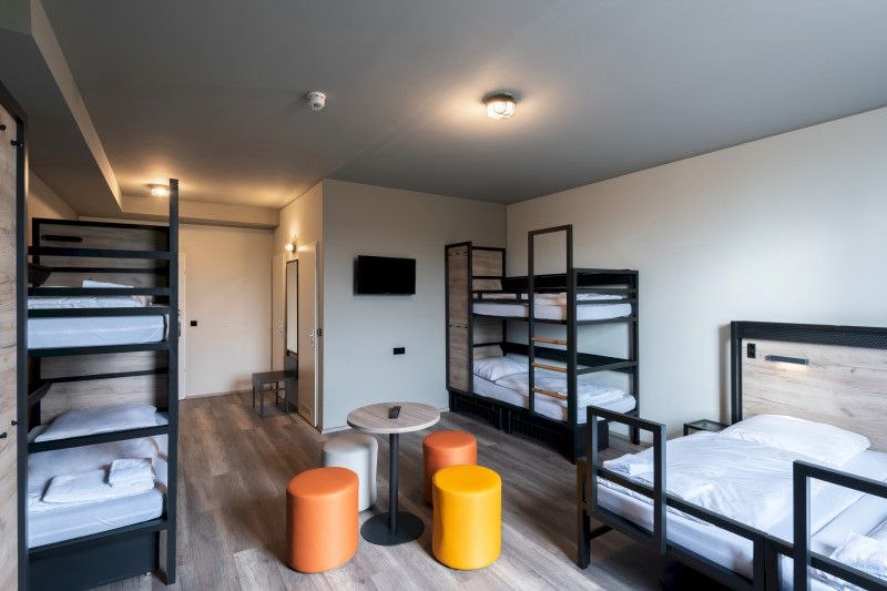 A modern family room with two bunk beds, a single bed, a small table and colorful stools. A television hangs on the wall., © A&O Hostels Marketing GmbH A modern family room with two bunk beds, a single bed, a small table and colorful stools. A television hangs on the wall., © A&O Hostels Marketing GmbH