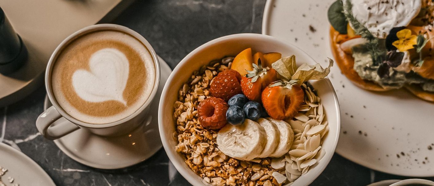 Ein Frühstückstisch mit einer Tasse Kaffee mit Herz-Latte-Art, einer Schüssel Müsli mit Früchten und einem Teller mit Waffeln und Avocado., © Fritz Stuttgart