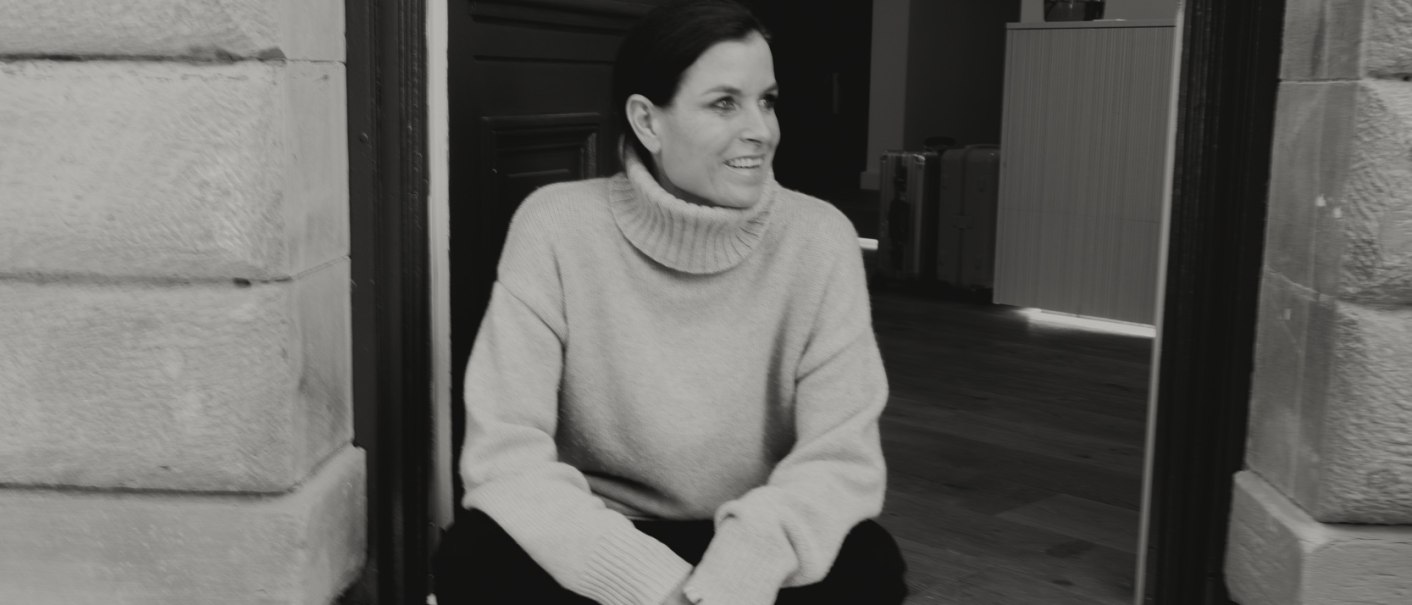 Eine Frau sitzt l&auml;chelnd auf einer T&uuml;rschwelle. Sie tr&auml;gt einen Rollkragenpullover und Sneakers. Der Hintergrund zeigt einen Innenraum., &copy; Peter Oliver Wolff / DAS EDITH