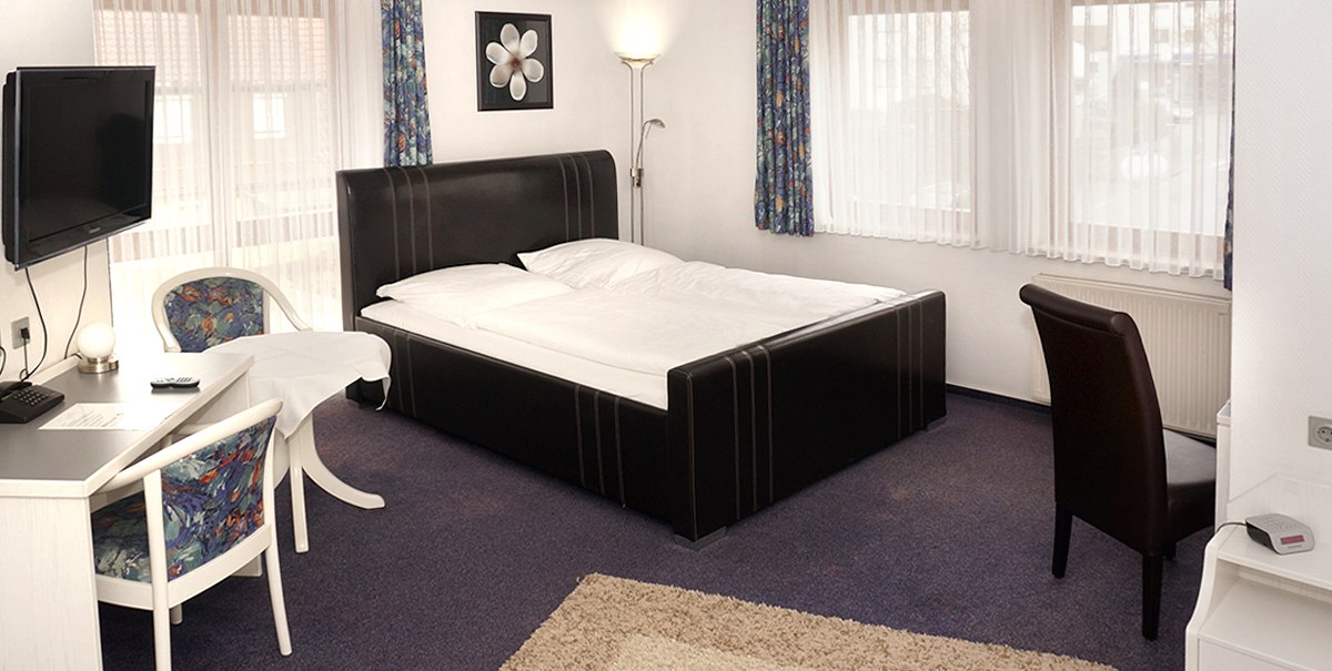 Helles Hotelzimmer mit Doppelbett, Schreibtisch, Stuhl und Fernseher. Große Fenster mit Vorhängen, Teppichboden und Wandbild., © TOMAS Helles Hotelzimmer mit Doppelbett, Schreibtisch, Stuhl und Fernseher. Große Fenster mit Vorhängen, Teppichboden und Wandbild., © TOMAS