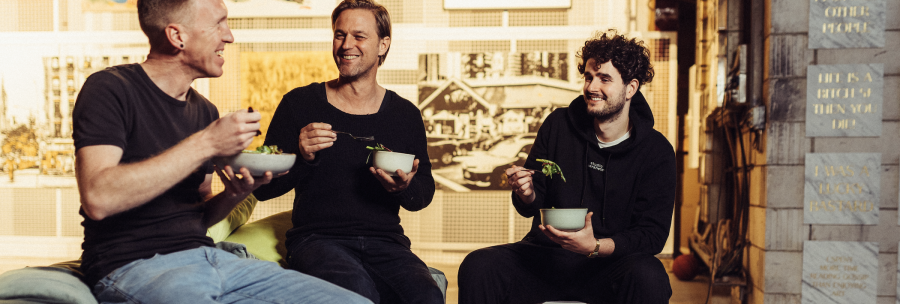 Three men sit together laughing and eating salad in a cozy, artistically designed room., © Vhy! GmbH Three men sit together laughing and eating salad in a cozy, artistically designed room., © Vhy! GmbH