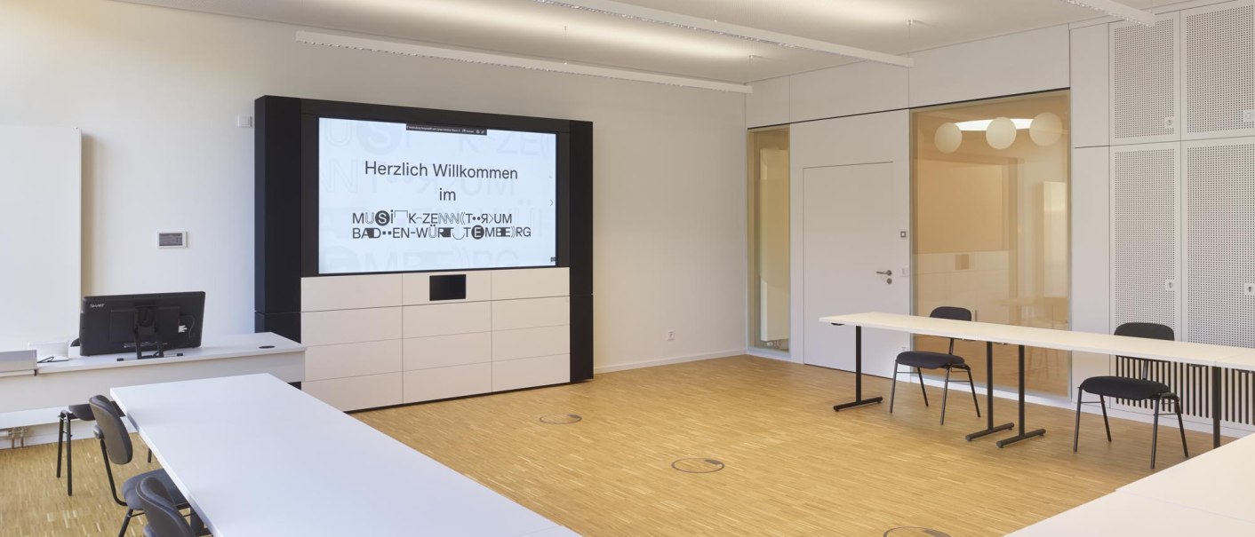 A modern seminar room with white tables and chairs, a large screen displaying a welcome message, wooden floors, and a glass wall., &copy; TOMAS