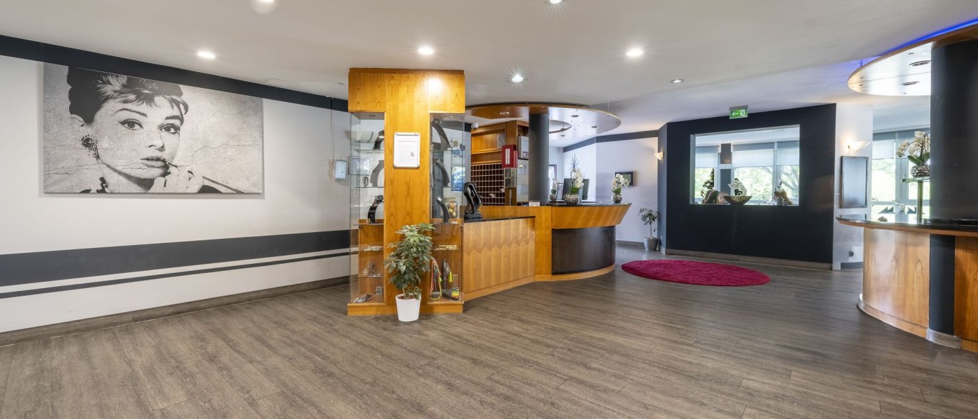 Modern reception lobby with wooden furniture, a large black and white portrait on the wall and a red carpet on the floor., © Plaza INN Stuttgart-Filderstadt Modern reception lobby with wooden furniture, a large black and white portrait on the wall and a red carpet on the floor., © Plaza INN Stuttgart-Filderstadt