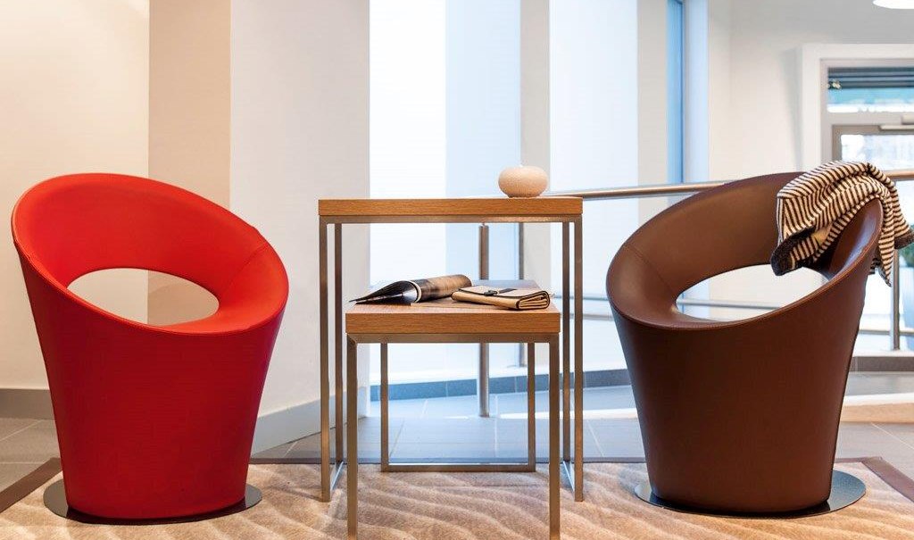 Zwei moderne Sessel, einer rot, einer braun, stehen in einer Lobby. Dazwischen ein kleiner Tisch mit Zeitschriften und Dekoration., © TOMAS Zwei moderne Sessel, einer rot, einer braun, stehen in einer Lobby. Dazwischen ein kleiner Tisch mit Zeitschriften und Dekoration., © TOMAS