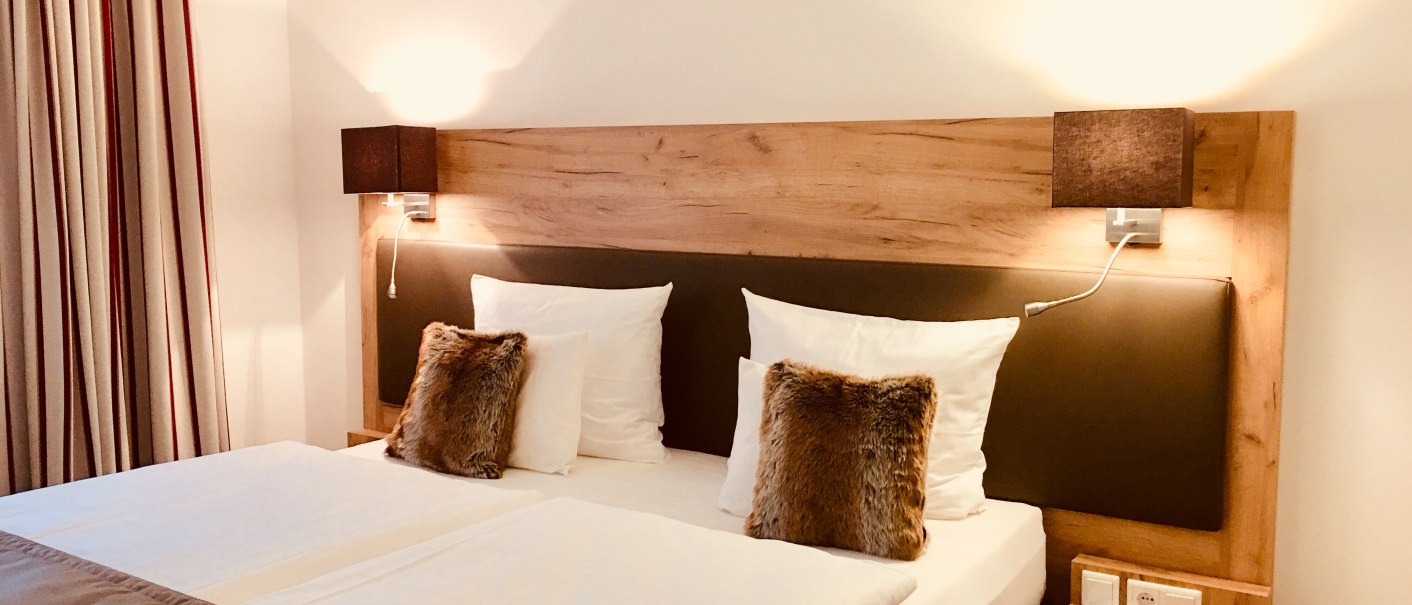 Modernes Hotelzimmer mit Doppelbett, Holzkopfteil, zwei Kissen und Nachttischlampen. Vorhänge an der Seite, gemütliche Atmosphäre., © TOMAS