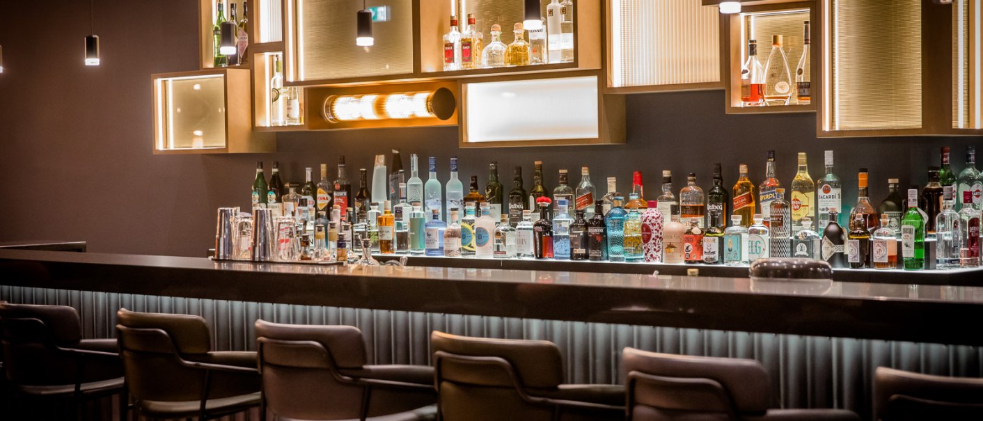 Elegante Bar mit stilvoller Beleuchtung, Flaschen und Gläsern auf der Theke. Moderne Hocker stehen bereit, die Atmosphäre ist gediegen und einladend., © TOMAS