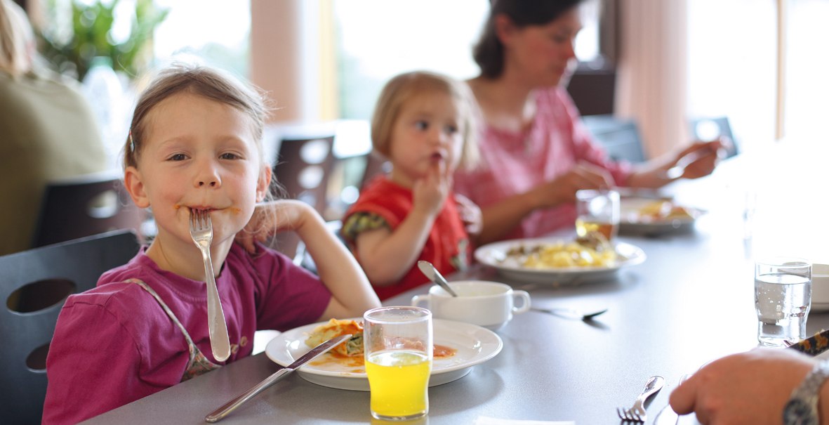 Two children and a woman are sitting at a table in a dining room, eating. One of the children has a fork in his mouth. There are plates and glasses on the table., © DJH Landesverband Baden-Württemberg e.V. Two children and a woman are sitting at a table in a dining room, eating. One of the children has a fork in his mouth. There are plates and glasses on the table., © DJH Landesverband Baden-Württemberg e.V.