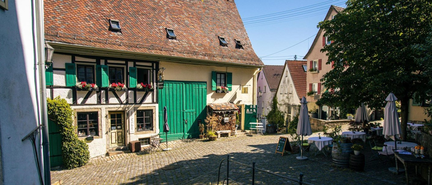 An idyllic courtyard in Nürtingen's old town with a half-timbered house, green shutters and cozy seating under a tree., © Stuttgart-Marketing GmbH, Sarah Schmid An idyllic courtyard in Nürtingen's old town with a half-timbered house, green shutters and cozy seating under a tree., © Stuttgart-Marketing GmbH, Sarah Schmid