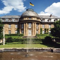 Die Villa Reitzenstein mit imposanter Fassade, einem Brunnen im Vordergrund und einer Flagge auf dem Dach, umgeben von Bäumen., © Staatsministerium Baden-Württemberg