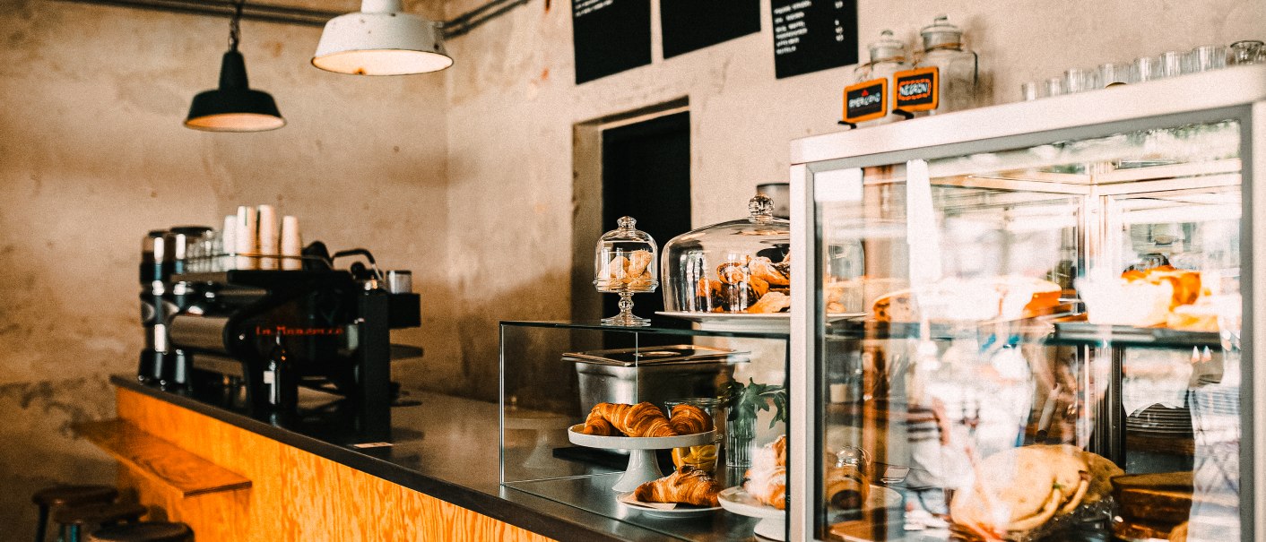 Ein gemütliches Café mit Holztresen, Kaffeemaschine und Gebäck in einer Glasvitrine. Hängelampen beleuchten den Raum., © Melanie Mearz Fotografie