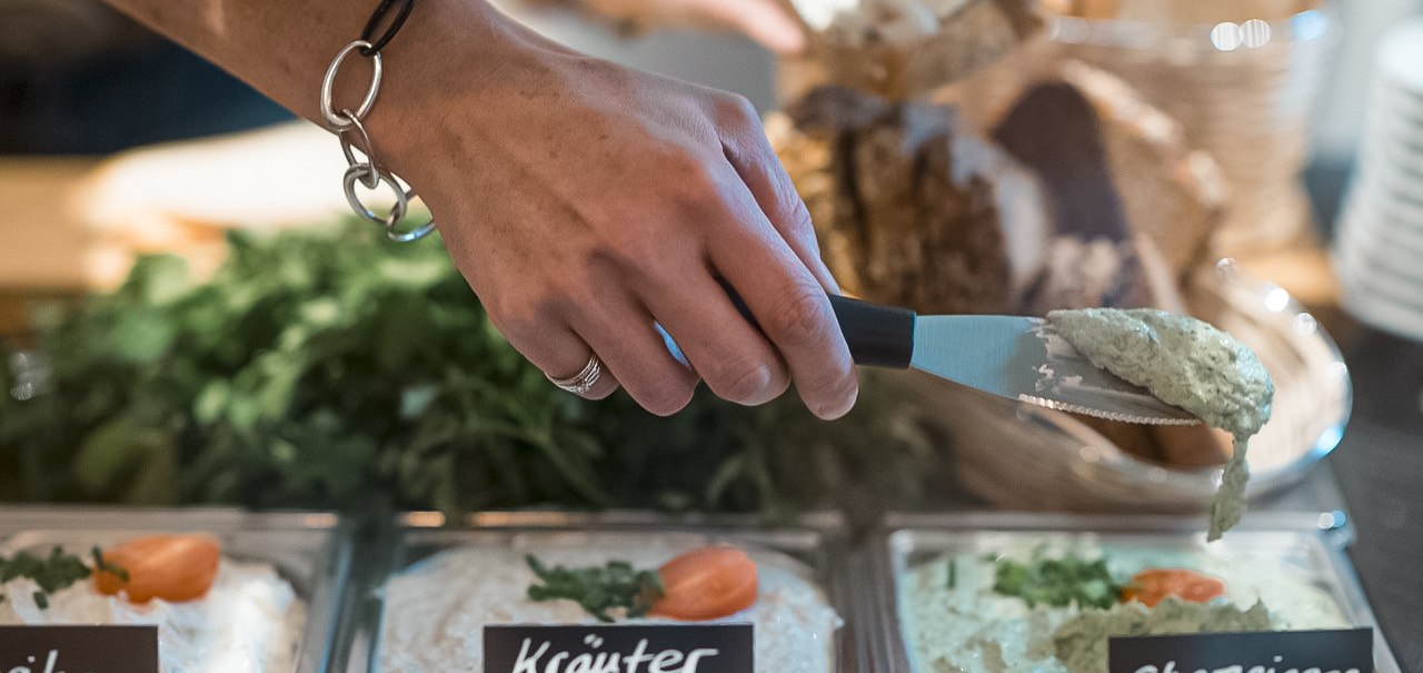 Eine Hand mit Armband nimmt Champignoncreme aus einer Schale. Daneben stehen Schalen mit Kräuterfrischkäse und Paprikacreme, im Hintergrund Brot., © Café Justus, Stuttgart Eine Hand mit Armband nimmt Champignoncreme aus einer Schale. Daneben stehen Schalen mit Kräuterfrischkäse und Paprikacreme, im Hintergrund Brot., © Café Justus, Stuttgart