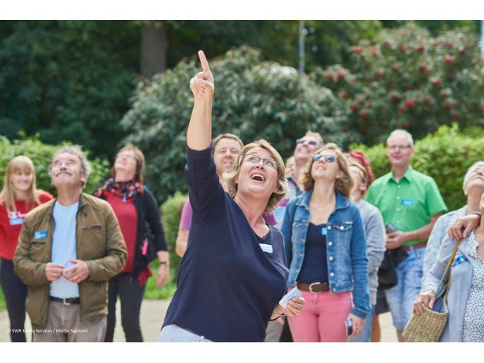 A woman points upwards while a group of people look in the same direction with interest. They are wearing name tags., &copy; SWR Media Services GmbH / Fernsehturm Stuttgart