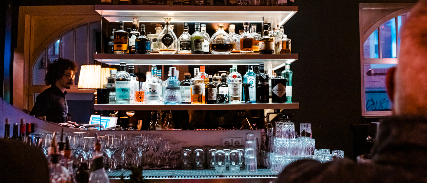 Bar mit beleuchteten Regalen voller Spirituosenflaschen, Gläsern und einem Barkeeper im Hintergrund. Gemütliche Atmosphäre mit gedämpftem Licht., © SMG, Sarah Schmid