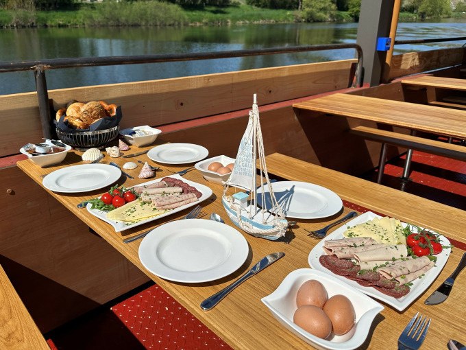 Fr&uuml;hst&uuml;ckstisch auf einem Schiff mit Wurst, K&auml;se, Eiern und Br&ouml;tchen. Im Hintergrund flie&szlig;t ein ruhiger Fluss vorbei., &copy; Cool-Tours StattReisen