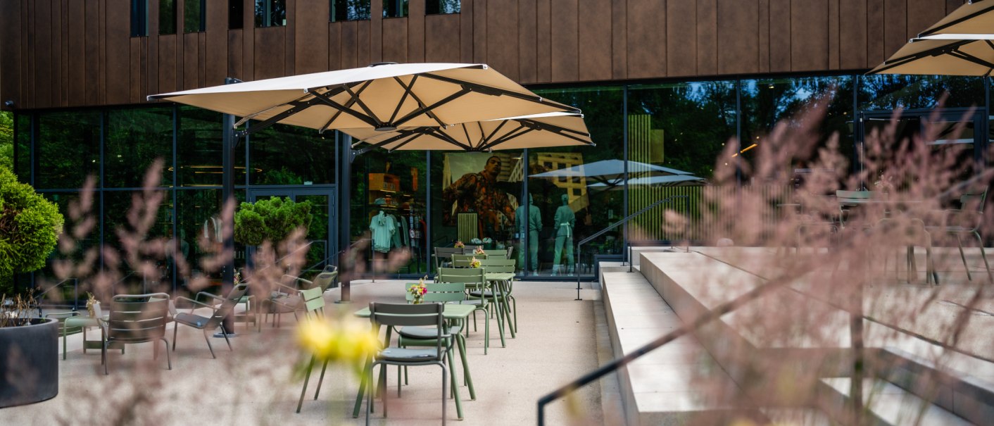 A modern outdoor café area with green tables and chairs under large umbrellas in front of a glass facade., © STIHL Markenwelt A modern outdoor café area with green tables and chairs under large umbrellas in front of a glass facade., © STIHL Markenwelt