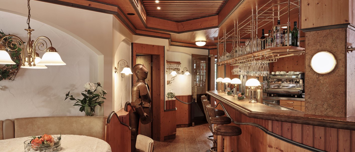 Gemütlicher Barbereich mit Holzeinrichtung, Barhockern und einer Ritterrüstung. Ein runder Tisch mit Blumenarrangement steht im Vordergrund., © Badhotel Restaurant Stauferland