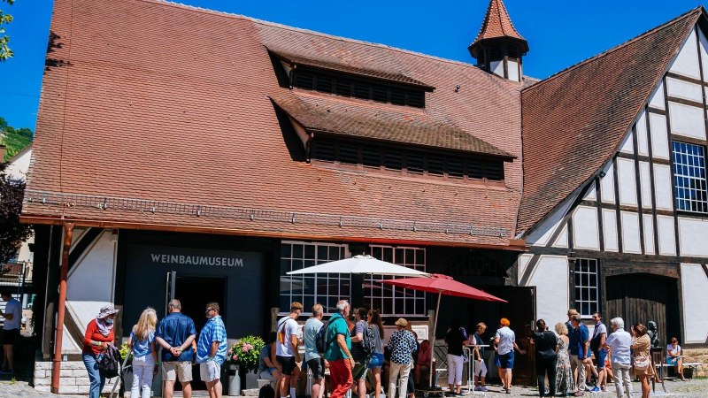 Besucher stehen vor dem Weinbaumuseum in Uhlbach. Das Fachwerkgebäude hat ein rotes Ziegeldach. Im Hintergrund sind Weinberge zu sehen., © Stuttgart-Marketing GmbH, Thomas Niedermüller