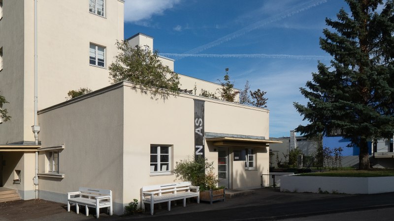 A beige architecture gallery with a flat roof, benches, and a large conifer tree against a blue sky., &copy; Thomas F&uuml;tterer