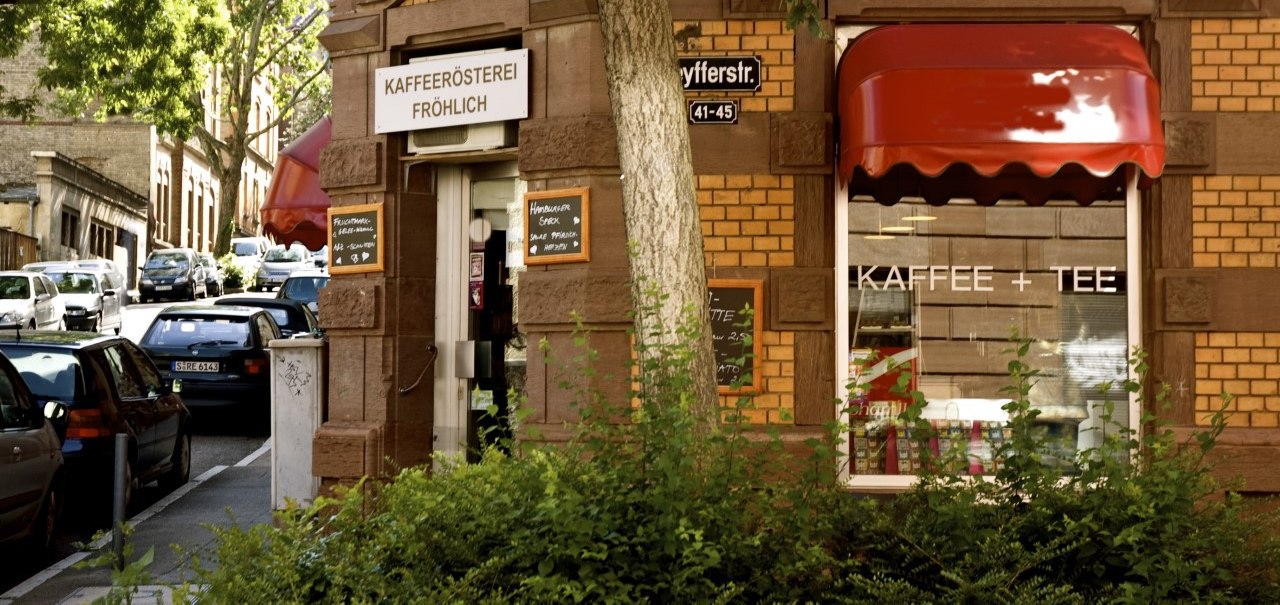 Entrance to the Fröhlich coffee roastery on a street corner with a red awning and signs. Cars are parked along the street, trees provide shade., © Kaffeerösterei Fröhlich