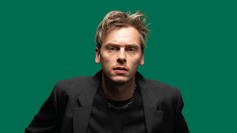 Ein Mann mit blondem Haar und schwarzem Mantel blickt ernst in die Kamera, vor einem grünen Hintergrund., © Württembergische Staatstheater Stuttgart Ein Mann mit blondem Haar und schwarzem Mantel blickt ernst in die Kamera, vor einem grünen Hintergrund., © Württembergische Staatstheater Stuttgart