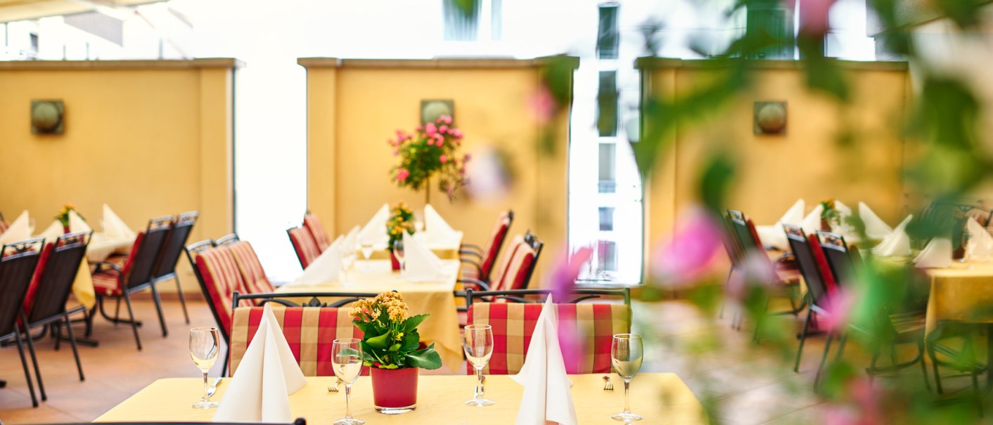 Elegante Terrasse mit gedeckten Tischen, Blumenarrangements und rot-gemusterten Stühlen unter einem Sonnendach., © Hirsch Hotel
