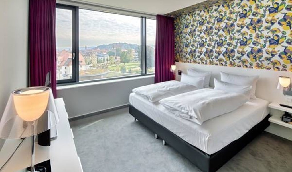 Modern hotel room with double bed, colorful wallpaper and large window with city view. Purple curtains and stylish lamps complete the design., &copy; Hotel am Remspark