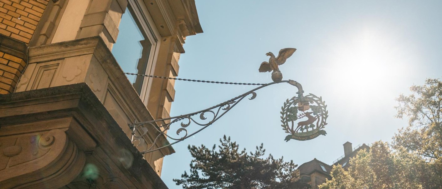 Ein Metallschild mit einem Adler und einem Hirsch hängt an einem Gebäude. Die Sonne scheint hell im Hintergrund., © SMG, Sarah Schmid