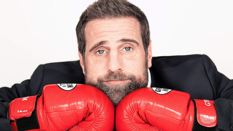 Ein Mann mit Bart stützt seinen Kopf auf rote Boxhandschuhe und schaut direkt in die Kamera. Er trägt einen dunklen Anzug., © Theaterhaus Stuttgart e.V. Ein Mann mit Bart stützt seinen Kopf auf rote Boxhandschuhe und schaut direkt in die Kamera. Er trägt einen dunklen Anzug., © Theaterhaus Stuttgart e.V.