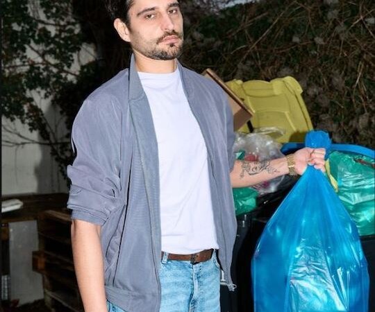 Ein Mann in Jeans und grauer Jacke h&auml;lt einen blauen M&uuml;llsack vor M&uuml;lltonnen. Der Hintergrund zeigt eine bewachsene Wand., &copy; Rosenau Kultur e.V.