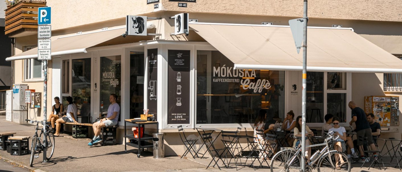 Café Mókuska an einer Straßenecke mit Außenbestuhlung und Gästen. Fahrräder sind geparkt, und es gibt Straßenschilder und einen Sonnenschutz., © SMG, Sarah Schmid